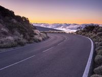 an empty winding road on a mountain with the sun set above the mountains in the background