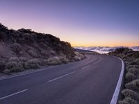 an empty winding road on a mountain with the sun set above the mountains in the background