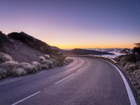 an empty winding road on a mountain with the sun set above the mountains in the background