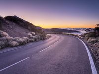 an empty winding road on a mountain with the sun set above the mountains in the background