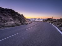 an empty winding road on a mountain with the sun set above the mountains in the background