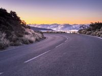 an empty winding road on a mountain with the sun set above the mountains in the background