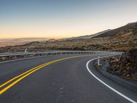 an empty road that is surrounded by a hill area at sunset with yellow lines on the side