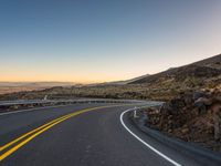 an empty road that is surrounded by a hill area at sunset with yellow lines on the side