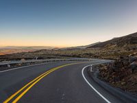 an empty road that is surrounded by a hill area at sunset with yellow lines on the side