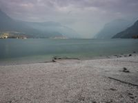 several people are walking along the shore on a gloomy day near mountains and water