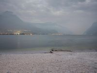 several people are walking along the shore on a gloomy day near mountains and water