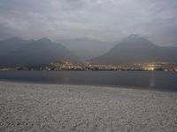 several people are walking along the shore on a gloomy day near mountains and water