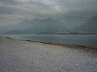 several people are walking along the shore on a gloomy day near mountains and water