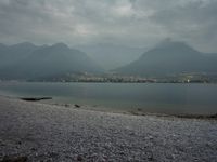 several people are walking along the shore on a gloomy day near mountains and water