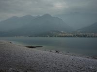 several people are walking along the shore on a gloomy day near mountains and water