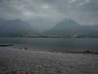 several people are walking along the shore on a gloomy day near mountains and water