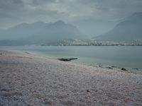 several people are walking along the shore on a gloomy day near mountains and water