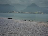 several people are walking along the shore on a gloomy day near mountains and water