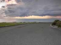 a stop sign sits next to a paved street near the ocean at sunset with a storm brewing