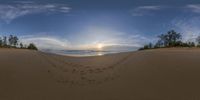 a view of the sun in the sky and clouds over the beach at sunset with footprints in the sand