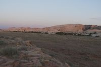 a hill in the middle of a desert area with rocks and vegetation on it, and another mountain in the distance