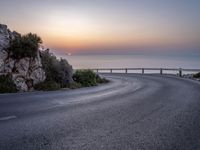 a curve road along the beach with the sunset on the horizon over the water behind it