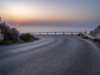 a curve road along the beach with the sunset on the horizon over the water behind it