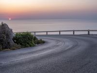 a curve road along the beach with the sunset on the horizon over the water behind it