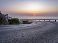 a curve road along the beach with the sunset on the horizon over the water behind it