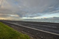 a view of a road next to the ocean and beach in the evening light with a sky and some clouds