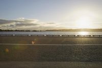 the sun shines over the water near a roadway with benches on the side of the road