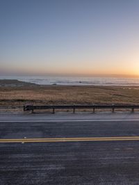 the road with yellow stripes is beside the ocean and some mountains at sunset in the distance