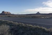 an empty dirt road leading to a desert scene, with tall trees, a mountain, and hills in the distance