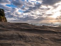 the sun is shining through dark clouds above a beach and ocean near a rocky shoreline