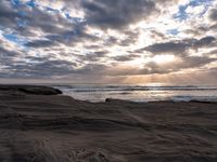 the sun is shining through dark clouds above a beach and ocean near a rocky shoreline