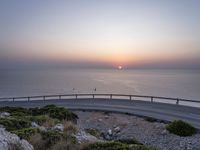 the sun setting over the ocean from the highway at the top of a hill with a bridge in foreground