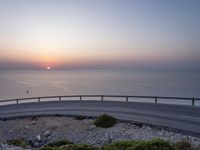 the sun setting over the ocean from the highway at the top of a hill with a bridge in foreground