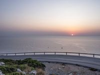 the sun setting over the ocean from the highway at the top of a hill with a bridge in foreground