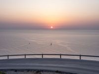 the sun setting over the ocean from the highway at the top of a hill with a bridge in foreground