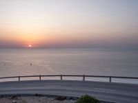 the sun setting over the ocean from the highway at the top of a hill with a bridge in foreground