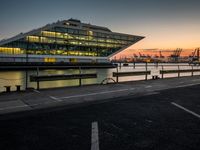 the sunset sets behind the waterfront where the city is setting at dusk, with cars parked along the shoreline and large glass windows on top of the building