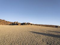 person with black backpack walking on large sandy field with hill in background near water, rocks, and sand