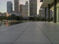 the walkway at the park leads to a lake and buildings in the background as evening falls