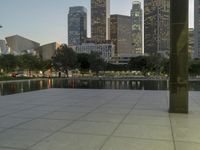 the walkway at the park leads to a lake and buildings in the background as evening falls