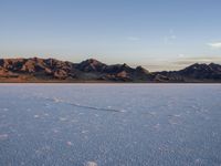 a vast open desert landscape with mountains in the background of blue sky and snow, on which is footprints that are drawn in the ground