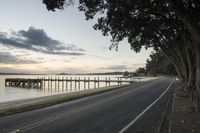 a road next to the water at sunset next to a dock and bridge in the distance