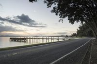 a road next to the water at sunset next to a dock and bridge in the distance