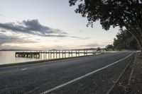 a road next to the water at sunset next to a dock and bridge in the distance