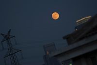 a large red full moon sits behind a skyscraper, high rise buildings and electricity lines