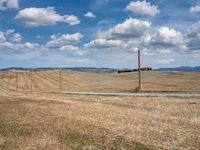 a field with dry grass and some power lines on it with clouds and a lone farm