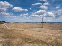 a field with dry grass and some power lines on it with clouds and a lone farm