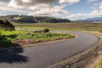 a paved country road winding off to the water and fields in the background with some trees