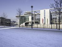 the bench is covered in snow and sits next to a building with glass windows, on the ground there is some tree