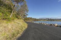 the side view of a paved road next to water with rocks around it and some trees in front of a large body of water